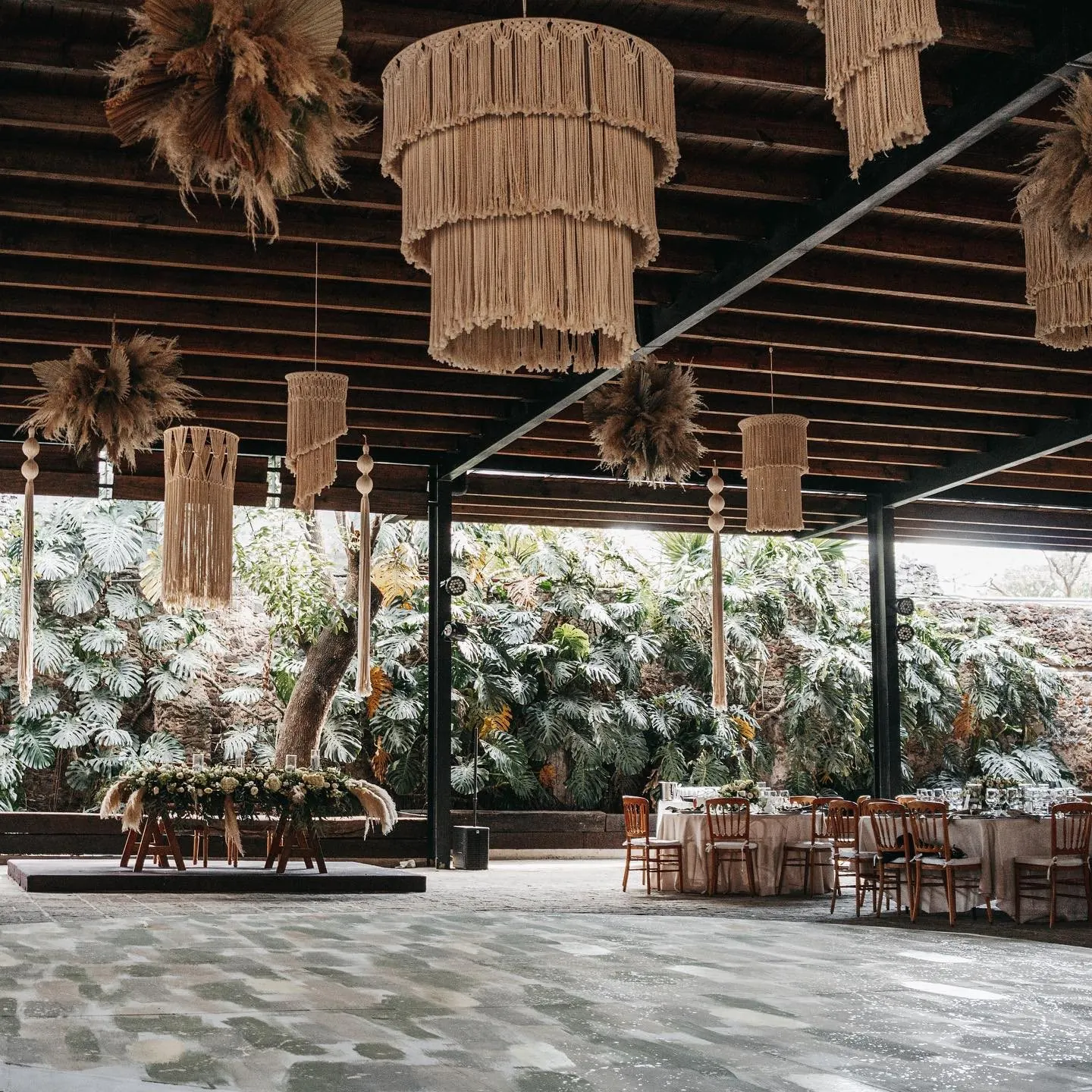 A reception hall with woven pampas-grass lamps hanging over long tables, garden visible behind open walls