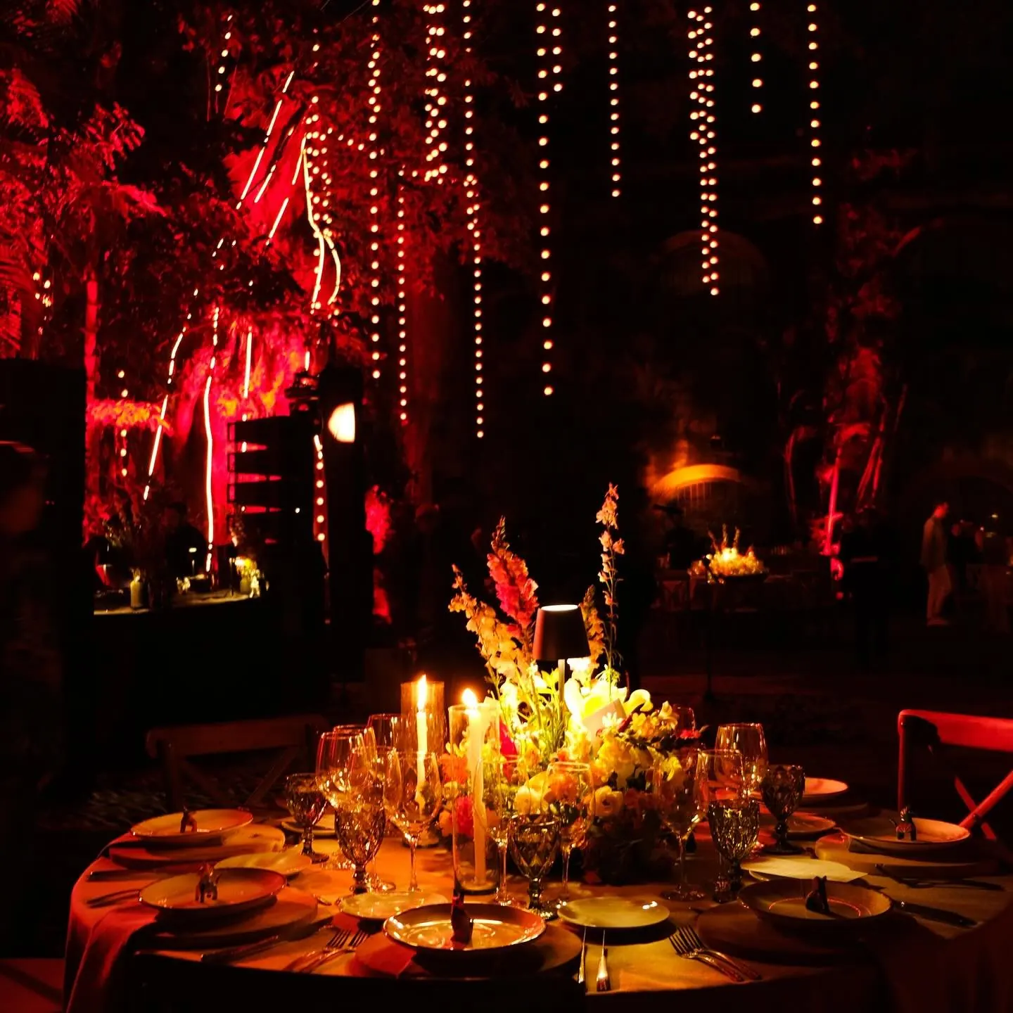 A long dinner table at night lit by candles and red light under hanging strands