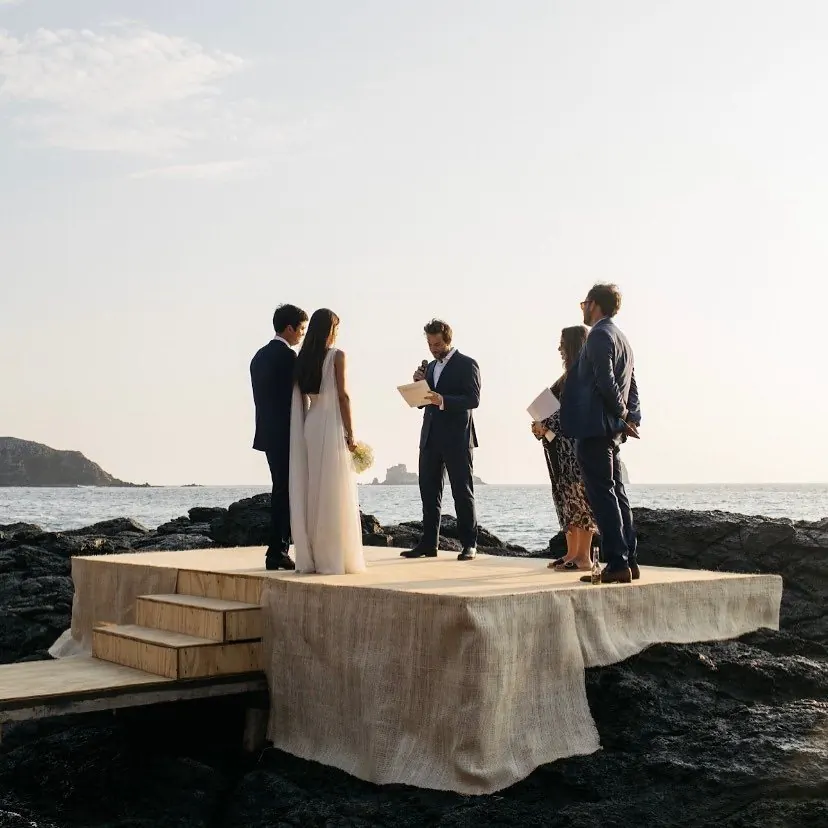 A couple and officiant on a draped platform on rocks above the sea at sunset